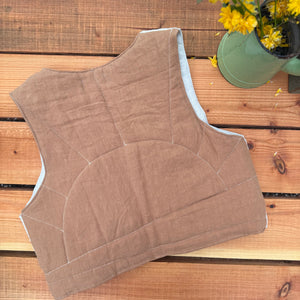 Brown quilted vest on a wooden surface with a potted plant in the background