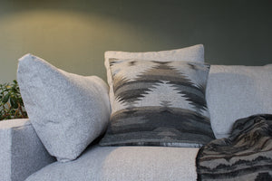 May include: A grey sofa with two grey throw cushions and a patterned throw cushion with a geometric design in shades of grey and black. A grey and black patterned throw blanket is draped over the sofa.