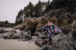 May include: A person wearing a blue and red hooded poncho with geometric patterns stands on a rocky beach. The person is looking out at the sea. The beach is surrounded by trees and rocks.