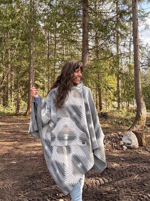 May include: A grey and white patterned poncho with a hood and a large pocket. The poncho is being worn by a person standing in a woodland setting.