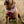 May include: A brown and tan dog with curly fur wearing a pink, red, orange, and purple bandana with a geometric pattern. The dog is looking up and has its mouth open.