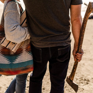 May include: A vintage axe with a wooden handle is held by a person. Another person holds a patterned tote bag and a matching blanket. A dog is in the background. The scene is set on a sandy beach.
