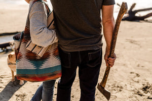 May include: A vintage axe with a wooden handle is held by a person. Another person holds a patterned tote bag and a matching blanket. A dog is in the background. The scene is set on a sandy beach.