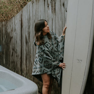 May include: A woman wearing a grey and black patterned poncho stands next to a white surfboard with the text "TORQ" printed on it. The surfboard is leaning against a wooden fence.