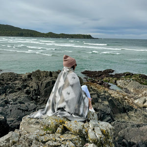 May include: A person wearing a pink beanie and a grey and white patterned blanket sits on a rocky cliff overlooking a body of water. The water is a light blue-green colour and there are waves crashing on the shore.