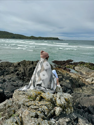 May include: A person wearing a pink beanie and a grey and white patterned blanket sits on a rocky cliff overlooking a body of water. The water is a light blue-green colour and there are waves crashing on the shore.